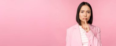 Sus, tabu kavramı. Asyalı iş kadınının portresi şşşş işareti, şşş işareti, parmak dudağa basma, pembe arka planda duran takım elbiseli