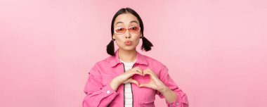 Güneş gözlüklü sevimli Koreli kadın model, kalbi gösteriyor, dikkat işareti, jestini seviyorum, pembe arka planda duruyor.