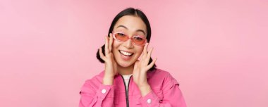 Güneş gözlüklü sevimli modern Japon kız, gülümsüyor ve mutlu görünüyor, pembe arka planda şık giyinmiş poz veriyor.