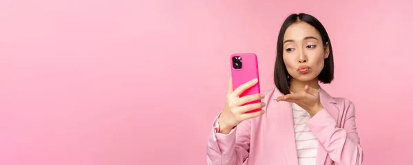 Стильная азиатская бизнесвумен, девушка в костюме делает селфи на смартфоне, видео-чат с приложением для мобильного телефона, позирует на фоне розовой студии — стоковое фото