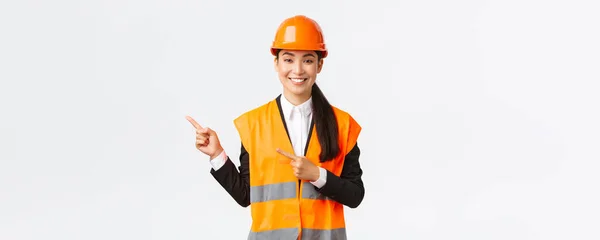Construção, construção e conceito industrial. Sorrindo asiático arquiteto feminino em capacete de segurança, roupas reflexivas apontando dedo canto superior esquerdo, mostrando projeto no local de trabalho, fundo branco — Fotografia de Stock