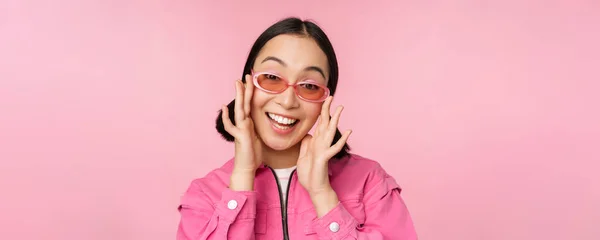 Симпатичная современная девушка в солнцезащитных очках, улыбающаяся и выглядящая счастливой, позирующая на розовом фоне в стильной одежде — стоковое фото