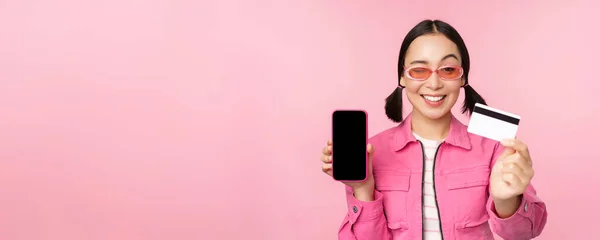 Bild av leende koreanska kvinna visar kreditkort och mobiltelefon skärm, smartphone applikationsgränssnitt, betala online, shopping kontaktlös, står över rosa bakgrund — Stockfoto