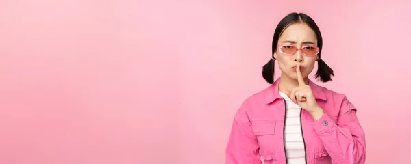 Moderní korejky dívka ve stylovém jarním oblečení, sluneční brýle, ukazující shush, ticho znamení, tisknout prst na rty, tabu gesto, stojící nad růžovým pozadím — Stock fotografie