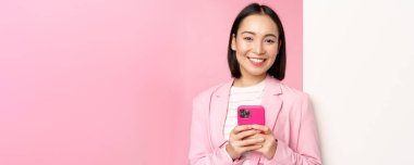 Takım elbiseli Koreli kadın girişimci görüntüsü, bilgi duvarının yanında duruyor, reklam güvertesinde, akıllı telefonu tutuyor ve gülümsüyor, pembe arka planda poz veriyor.