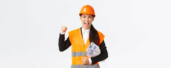 Succesvolle vrouwelijke Aziatische ingenieur, architect in veiligheidshelm en reflecterende jas dragen blauwdrukken van bouwproject en vuist pomp in viering, schreeuwen ja, winnende tender, witte achtergrond — Stockfoto