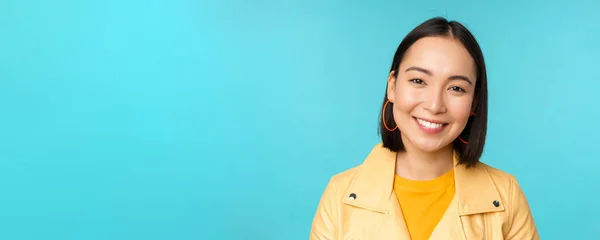 Close-up portret van glimlachende mooie aziatische vrouw met witte tanden, kijken blij op camera, poseren in gele jas over blauwe studio achtergrond — Stockfoto