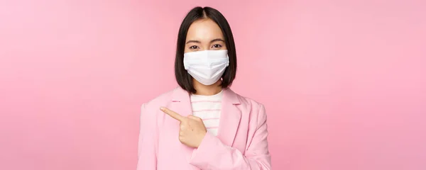 Портрет азійської бізнесменки в медичній масці та костюмі, вказівний палець зліва, показуючи рекламу, прапор компанії, студійний рожевий фон — стокове фото