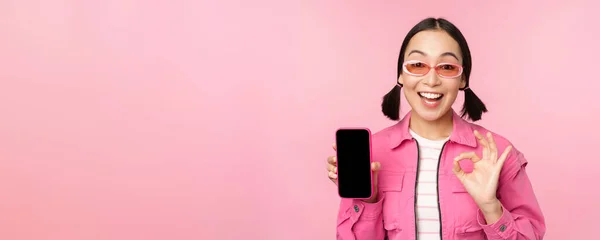 Enthousiaste jonge aziatische vrouw tonen oke, ok teken, glimlachen blij, mobiele telefoon scherm, smartphone applicatie, staande over roze achtergrond — Stockfoto