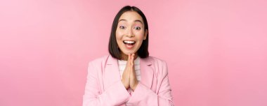 Mutlu Asyalı ofis kadınının portresi, takım elbiseli iş kadını seviniyor ve gülüyor, kazanıyor, kutluyor, amacına ulaşıyor ve seviniyor, pembe arka planda duruyor.