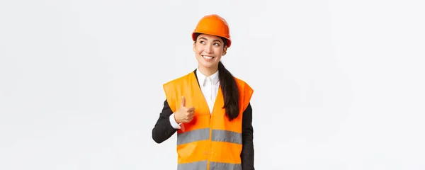 Щаслива усміхнена жінка-азіатка-архітектор задоволена будівельними роботами, стоячи в шоломі безпеки і відбиваючій куртці, дивлячись у верхній лівий кут і показуючи великі пальці на схвалення — стокове фото