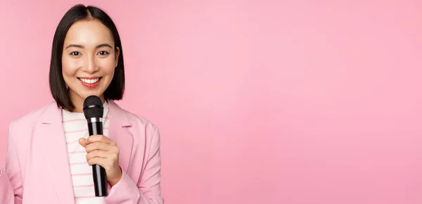 Ασιάτισσα επιχειρηματίας δίνοντας ομιλία, κρατώντας μικρόφωνο και χαμογελώντας, στέκεται στο κοστούμι πάνω από ροζ φόντο — Φωτογραφία Αρχείου