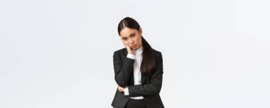 Annoyed and exhausted asian saleswoman looking unamused and bothered from under forehead, standing in black suit, leaning on palm and stare pissed-off, standing white background