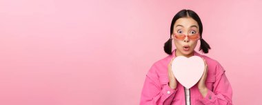 Güzel Asyalı kız mutlu gülümsüyor, kalp hediye kutusunu gösteriyor ve kameraya heyecanlı bakıyor, pembe romantik arka planda duruyor.
