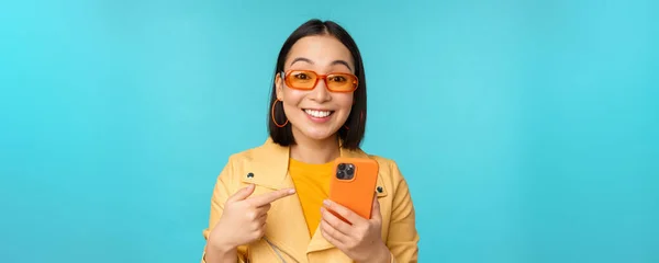 Entusiasta ragazza asiatica in occhiali da sole alla moda, puntando il dito verso smartphone, mostrando il telefono cellulare, in piedi su sfondo blu — Foto Stock