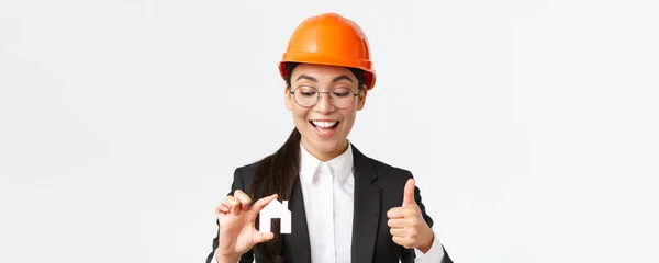 Gros plan de heureux heureux architecte asiatique femme, ingénieur de construction en casque et costume regardant miniature à la maison et montrant pouces vers le haut, garantir la qualité des travaux de réparation, fond blanc — Photo