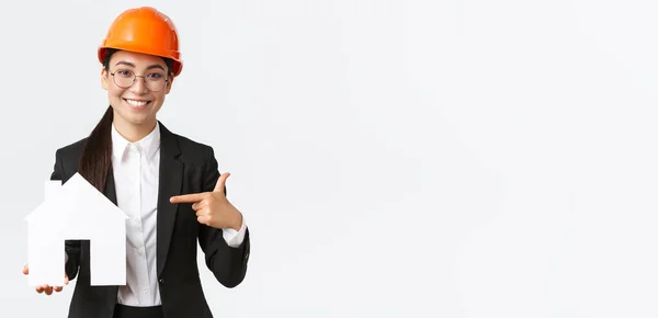 Retrato de jovem asiático engenheiro de construção feminino, arquiteto em capacete de segurança e terno de negócios apontando dedo em casa maket e sorrindo, promovendo o serviço da empresa de construção de casas — Fotografia de Stock
