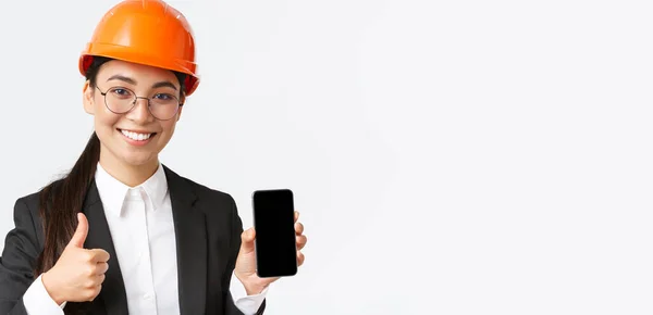 Zbliżenie profesjonalnej uśmiechniętej kobiety inżynier, kierownik budowy w garniturze biznesowym i kasku bezpieczeństwa, pokazując ekran smartfona i kciuki do góry w zatwierdzeniu, białe tło — Zdjęcie stockowe