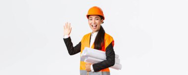 Dost canlısı, neşeli Asyalı kadın inşaat yöneticisi, güvenlik kaskı ve ceketli baş mimar, bina projesinin planları, el sallayarak merhaba demek, birini selamlamak,