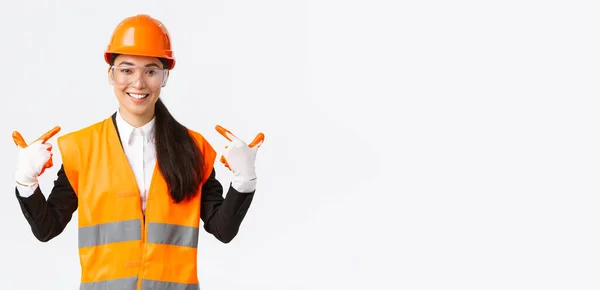 Sonriente confiada asiática jefa de ingeniería de construcción en casco de seguridad, guantes y gafas apuntándose a sí misma, notificar a seguir el protocolo seguro y usar ropa especial, fondo blanco — Foto de Stock