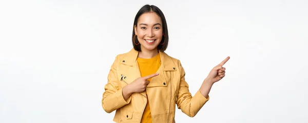 Mujer asiática feliz sonriendo, señalando con los dedos a la derecha, invitando a revisar la venta, mostrando banner publicitario o logotipo, de pie sobre fondo blanco —  Fotos de Stock