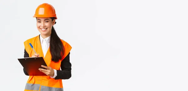 Uśmiechnięty zadowolony azjatycki inżynier budowlany prowadzi inspekcję w przedsiębiorstwie, nosi kask bezpieczeństwa i kurtkę odblaskową, zapisuje notatki i wygląda zadowolony wynik, białe tło — Zdjęcie stockowe