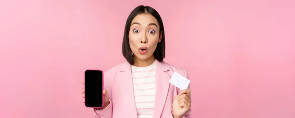 Entusiastiska asiatiska affärskvinna visar mobiltelefon skärm och kreditkort, ser förvånad över kameran, står över rosa bakgrund — Stockfoto