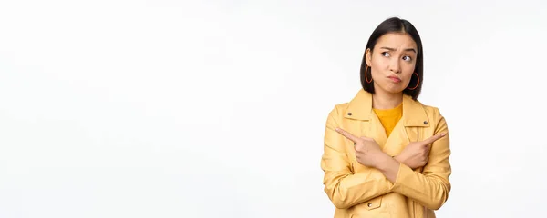 Image d'une fille asiatique indécise, pointant du doigt latéralement, pointant à gauche et à droite, choisissant une variante, décidant, debout sur fond blanc — Photo