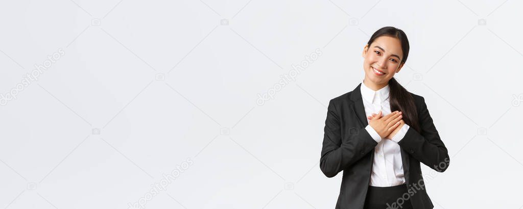 Smiling happy asian female entrepreneur receive praises or compliments, holding hands on heart, grinning and looking flattered, feeling thankful, standing white background delighted