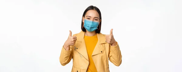 Mosolygó ázsiai nő orvosi maszk mutatja hüvelykujját felfelé, támogatást visel védőfelszerelés covid-19, coronavírus pandémia, fehér háttér — Stock Fotó