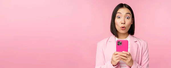 Portret van Aziatische zakenvrouw met een verrast gezicht, met behulp van smartphone app, het dragen van business suit. Koreaans meisje met mobiele telefoon en opgewonden gezicht expressie, roze achtergrond — Stockfoto