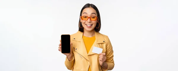 Nätshopping och människor koncept. Snygg asiatisk kvinna som visar mobiltelefon skärm och kreditkort, smartphone ansökan, stående över vit bakgrund — Stockfoto