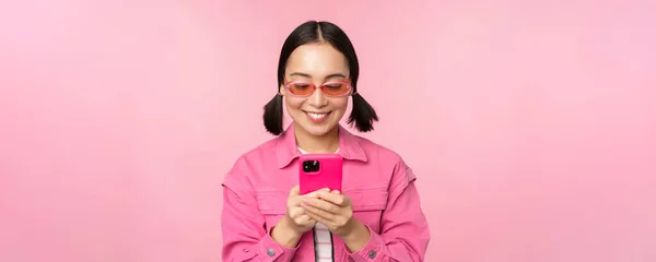 Porträtt av asiatisk flicka i solglasögon med hjälp av smartphone. Kvinna tittar på mobiltelefon, bläddrar i appen, står över rosa bakgrund — Stockfoto