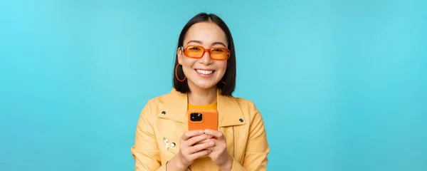 Retrato de mujer asiática entusiasta en gafas de sol, usando teléfono móvil, sonriendo y riendo, mirando feliz, sosteniendo teléfono inteligente, de pie sobre fondo azul — Foto de Stock