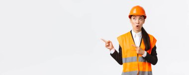 İnşaat, inşaat ve endüstriyel konsept. İlgi çekici ve şaşırmış kadın mühendis, miğfer ve yansıtıcı giysiler giyen Asyalı mimar, parmaklarını sol üst köşeyi göstererek 
