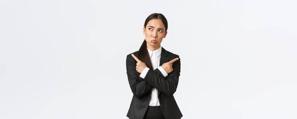 Озадаченная симпатичная азиатка-бизнесмен в черном костюме с трудом делает выбор, стоит нерешительно, указывает пальцем в сторону и выглядит сомнительно, думая о том, какое решение принять, стоя на белом фоне