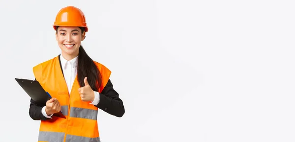 Satisfait ingénieur de construction asiatique satisfait des résultats de l'inspection, portant des vêtements de sécurité et un casque à la zone du bâtiment, note et montrer pouces levés dans l'approbation — Photo