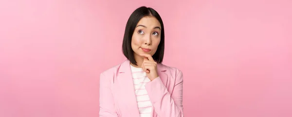 Imagem de mulher de negócios asiática em pé em pose de pensamento, brainstoming, vestindo terno. Vendedora coreana, empresária posando contra fundo rosa — Fotografia de Stock