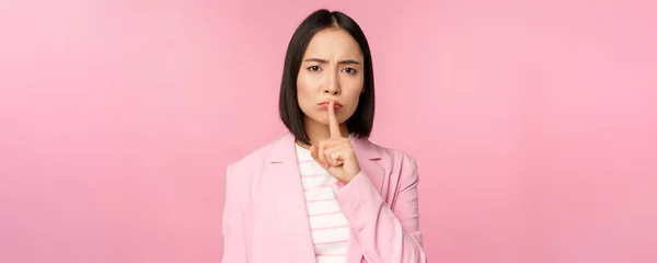 Silenzio, concetto tabù. Ritratto di donna d'affari asiatica che mostra gesto di zittire, segno di shhh, premere dito sulle labbra, in piedi su sfondo rosa in abito — Foto Stock