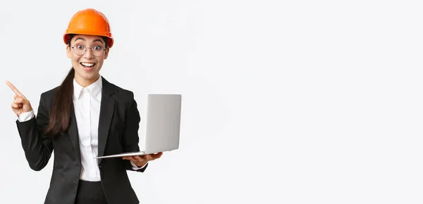 Excited happy asian female engineer, industrial woman in safety helmet and business suit, showing presentation, pointing finger at graph or chart and holding laptop computer, smiling amazed — Stock Photo, Image