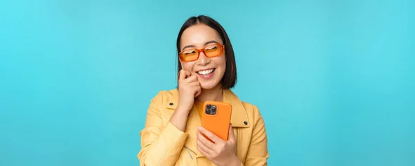 Felice ragazza asiatica elegante utilizzando smartphone e ridere, sorridendo alla fotocamera, in piedi su sfondo blu — Foto Stock