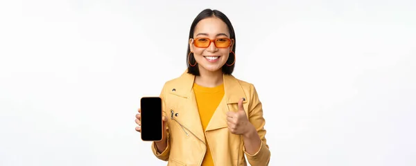 Красивая корейская девушка, азиатка в солнцезащитных очках, показывает интерфейс приложения смартфона, большие пальцы вверх, рекомендуя мобильное приложение, белый фон — стоковое фото