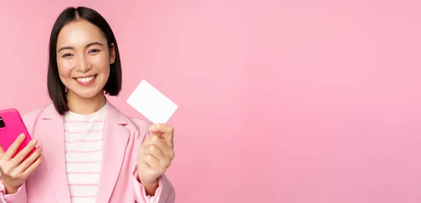 Bild av leende glad asiatisk affärskvinna visar kreditkort, betala online på smartphone ansökan, beställa med mobil phon, står mot rosa bakgrund — Stockfoto