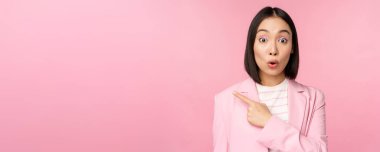 Şaşırmış yüzlü bir iş kadını portresi, sol eli işaret eden, smth 'i ilginç gösteren, ofis elbisesinin içinde pembe arka planda duran