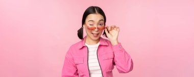 Göz giysisi reklamı. Modern Asyalı zarif bir kız güneş gözlüğüne dokunur, pembe giyer, stüdyo arka planında poz verir. Boşluğu kopyala