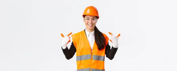 Sonriente confiada asiática jefa de ingeniería de construcción en casco de seguridad, guantes y gafas apuntándose a sí misma, notificar a seguir el protocolo seguro y usar ropa especial, fondo blanco — Foto de Stock