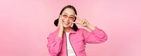 Portrét kawaii asijské dívky v slunečních brýlích, ukazující mír, V-znamení v blízkosti oka a usmívá roztomilý na kameru, pózování v módní oblečení proti růžovému pozadí — Stock fotografie