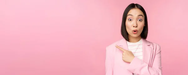Portrait de femme d'affaires avec visage surpris, doigt pointant vers la gauche, montrant smth intéressant, debout sur fond rose en costume de bureau — Photo