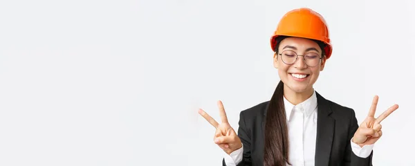Közelkép vidám sikeres női ázsiai mérnök, építőipari építész biztonsági sisak és üzleti ruha mutatja békejelek és mosolygós kawaii, álló fehér háttér — Stock Fotó