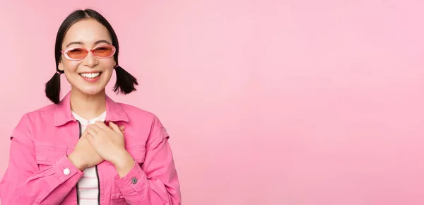 Sorrindo jovem mulher asiática em óculos de sol olhando grato, de mãos dadas no coração, lisonjeado, apreciar smth, de pé sobre fundo rosa — Fotografia de Stock